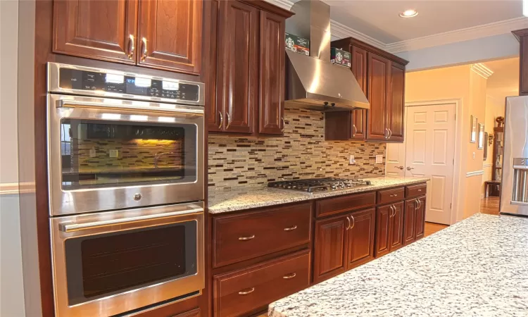 Kitchen with light stone counters, stainless steel appliances, exhaust hood, tasteful backsplash, and crown molding