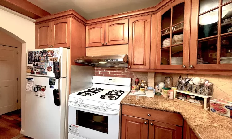 Kitchen with white appliances, backsplash, light stone counters, and under cabinet range hood