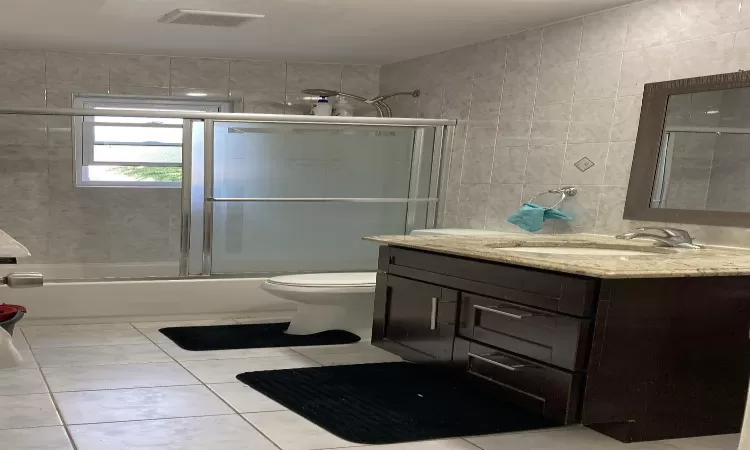 Bathroom featuring visible vents, toilet, vanity, combined bath / shower with glass door, and tile walls