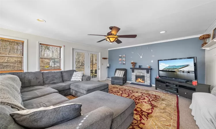 Carpeted living room with a stone fireplace, baseboards, crown molding, and a ceiling fan