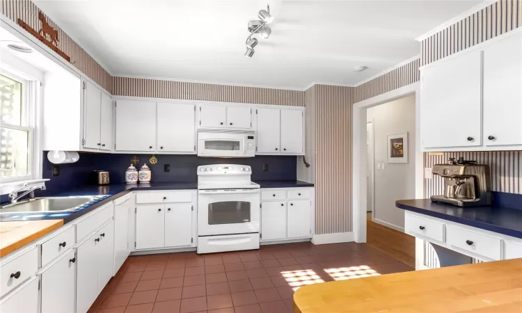 Kitchen with wallpapered walls, dark tile patterned flooring, white cabinets, white appliances, and a sink