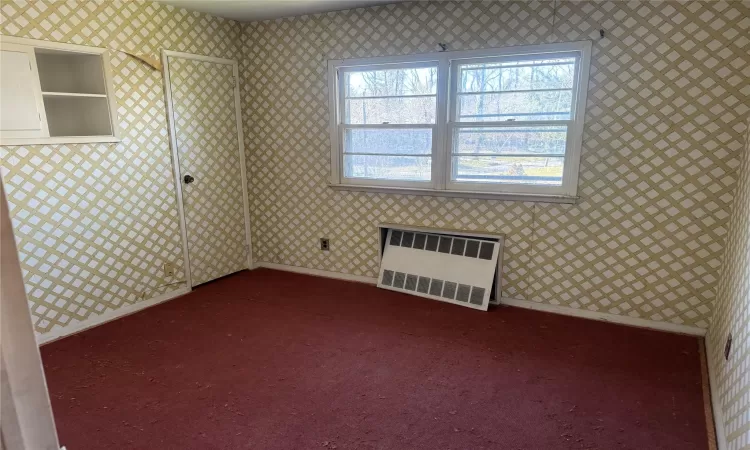 Empty room featuring radiator, wallpapered walls, baseboards, and dark colored carpet