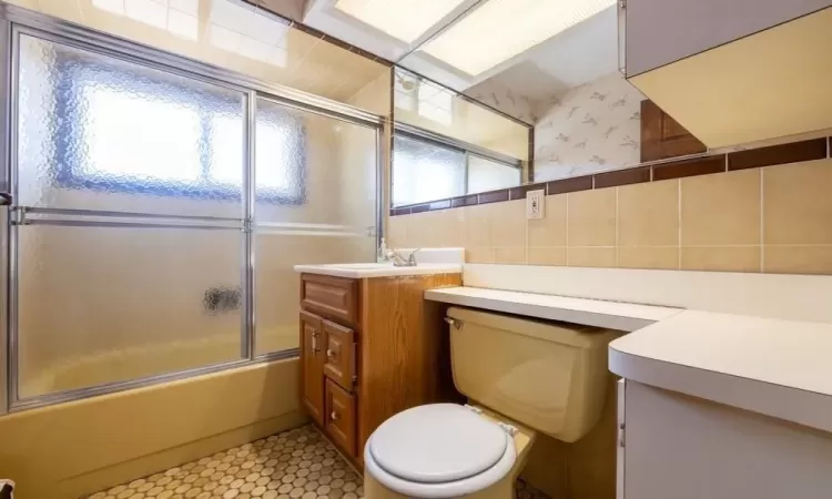 Bathroom with wallpapered walls, toilet, vanity, shower / bath combination with glass door, and tile walls