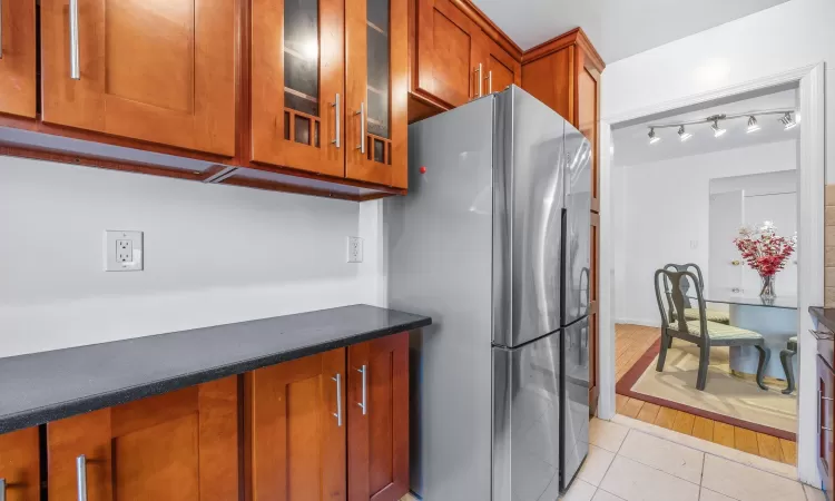 Kitchen with dark countertops, freestanding refrigerator, light tile patterned flooring, brown cabinetry, and glass insert cabinets