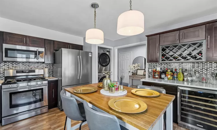 Kitchen with light countertops, wine cooler, dark brown cabinets, stacked washer and clothes dryer, and appliances with stainless steel finishes