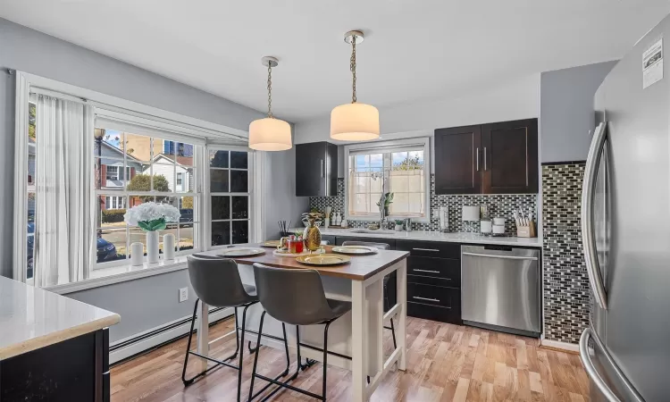 Kitchen with light wood finished floors, a baseboard radiator, stainless steel appliances, hanging light fixtures, and backsplash