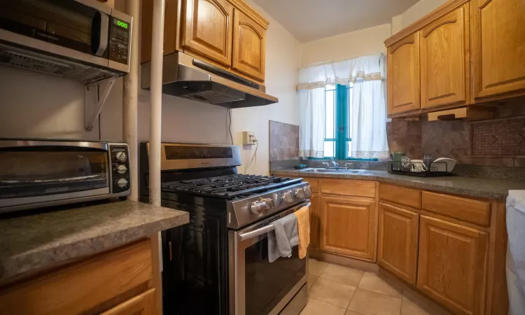 Kitchen with light tile patterned floors, stainless steel appliances, under cabinet range hood, dark countertops, and tasteful backsplash